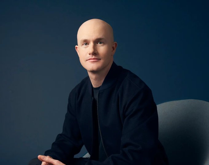 Brian Armstrong, the bald CEO of Coinbase, sits thoughtfully in a dark bomber jacket against a deep blue background, looking off-camera with a calm expression. The image accompanies a warning from him that his social media activity should not be interpreted as investment advice.