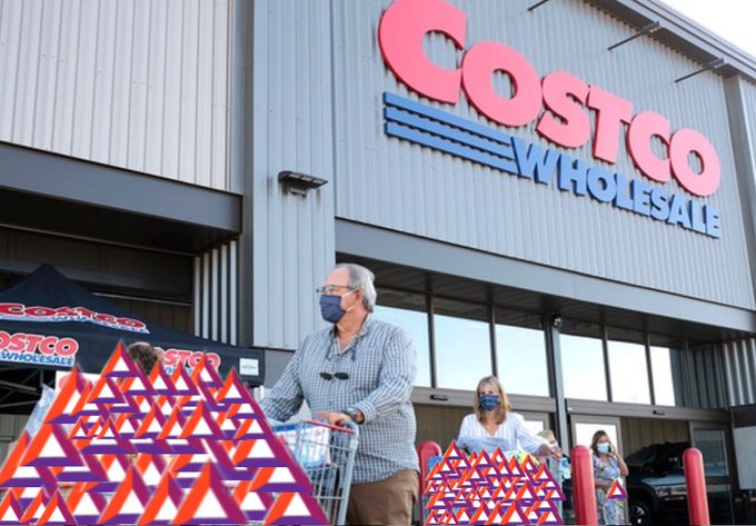 An outdoor photograph of a Costco Wholesale storefront with shoppers wearing masks pushing shopping carts, with red and orange geometric memorial structures in the foreground.
