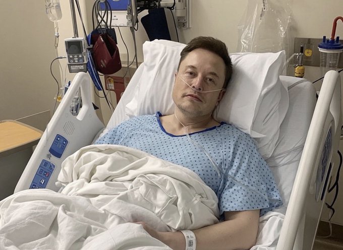 Elon Musk lies in a hospital bed wearing a blue gown and nasal cannula, appearing weary and defeated, symbolizing the painful financial consequences of the $LCID vs $SNDK investment comparison. The post contrasts buying Lucid at its peak of $21 versus SanDisk at $47, illustrating how the "wrong" choice led to staggering losses for investors.