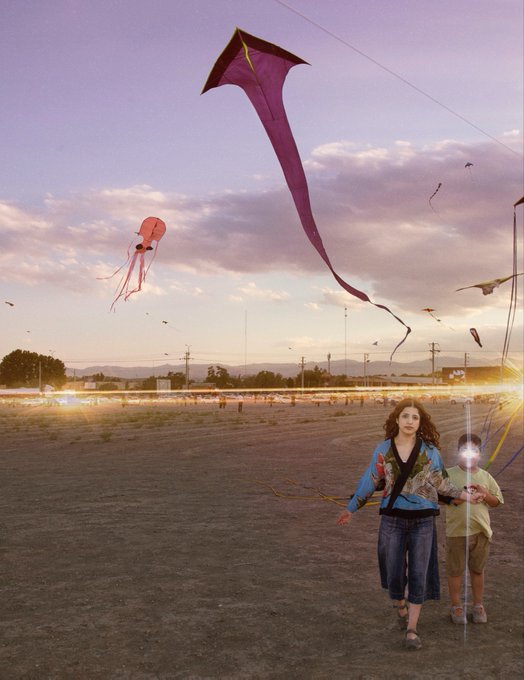 Two people walk across a sandy field at sunset while multiple kites fly in the pastel-colored sky above them.