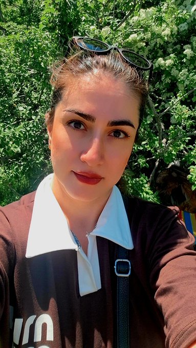A selfie of a young woman with sunglasses perched on her head, wearing a brown and white collared shirt, posing in front of green foliage.