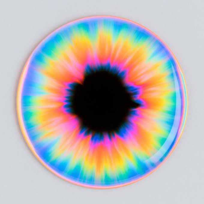 A circular sticker or pin featuring a vibrant rainbow tie-dye iris design with a black heart-shaped pupil in the center.