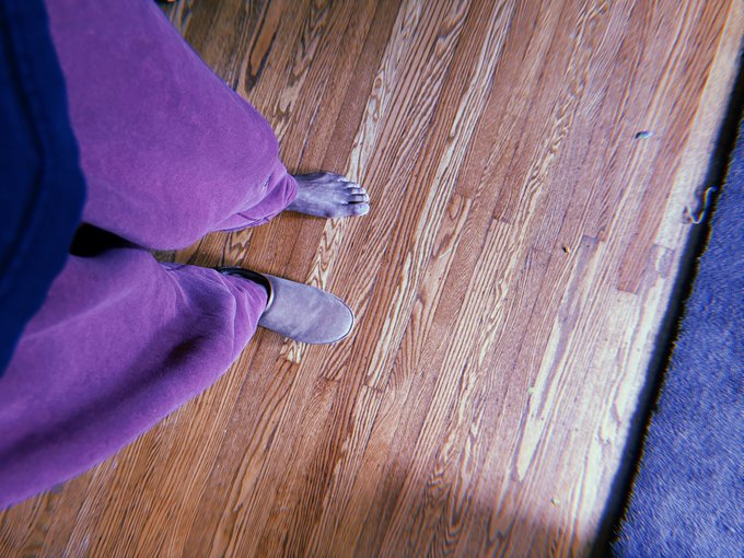 A downward view of a person wearing purple sweatpants and slippers standing on a wooden plank floor next to a blue rug.