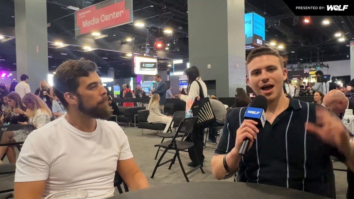 The image shows two men sitting across from each other, one of them holding a microphone while they are talking. They appear to be discussing something related to media or technology. In the background, there are several people seated at tables with chairs around them, suggesting that this could be an event or gathering taking place in a large room.

The image also contains text elements such as "WOLF" and "Immunefi", which may provide additional context about the event or the individuals involved.