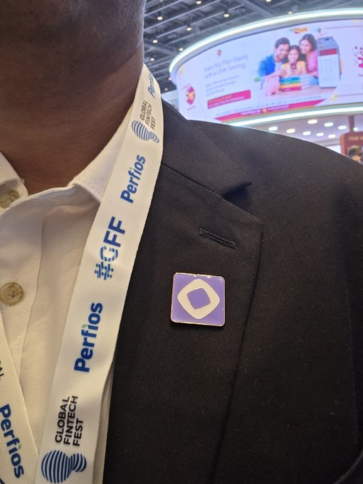 A man in a dark suit wears a 'Perifos' Global Fintech Fest lanyard and a square purple and white badge on his lapel at what appears to be a convention.