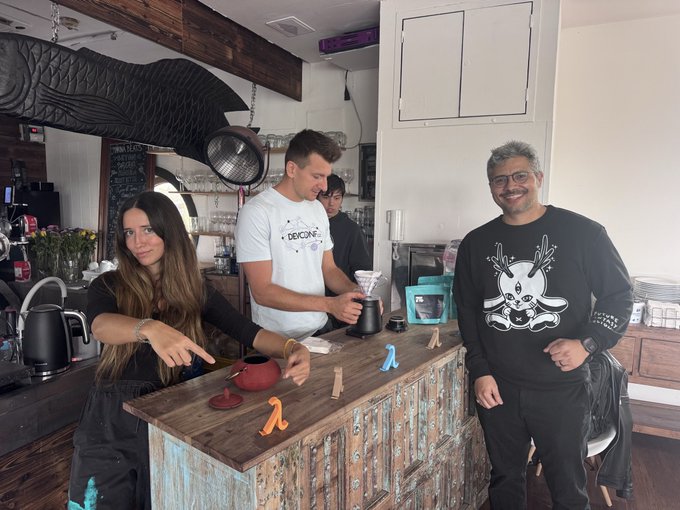 Three people standing behind a wooden counter in a cafe or coffee shop setting, with one man wearing a black sweatshirt featuring a white bunny skull logo.