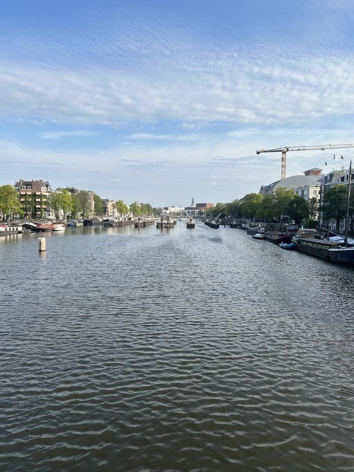 A tranquil canal scene under a bright blue sky with wispy clouds, featuring houseboats lining both banks and a construction crane visible in the distance. The image complements the "Good morning" greeting with its peaceful, sunlit waterways and crisp morning atmosphere.