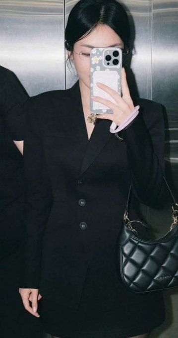 A woman with black hair takes a mirror selfie in an elevator while wearing a black blazer dress, carrying a quilted black handbag and holding a phone with a daisy-patterned case.