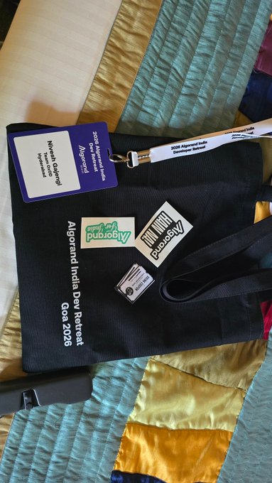 The image features a black bag with various stickers, including one that says "Algofam" and another that reads "Algofam I am here in goa for the Algofam Dev Retreat by @Algo_Bharat Super Hyped to See fellow devs build something cool By the beach !!!". The bag is placed on a bed with a colorful quilt, which adds an interesting visual element to the scene.