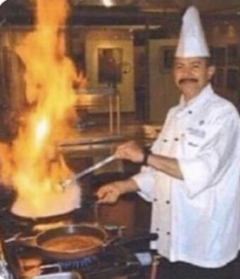 A chef in traditional white uniform and tall toque hat smiles while cooking at a stove, with a large flame flaring up from a pan and holding utensils.