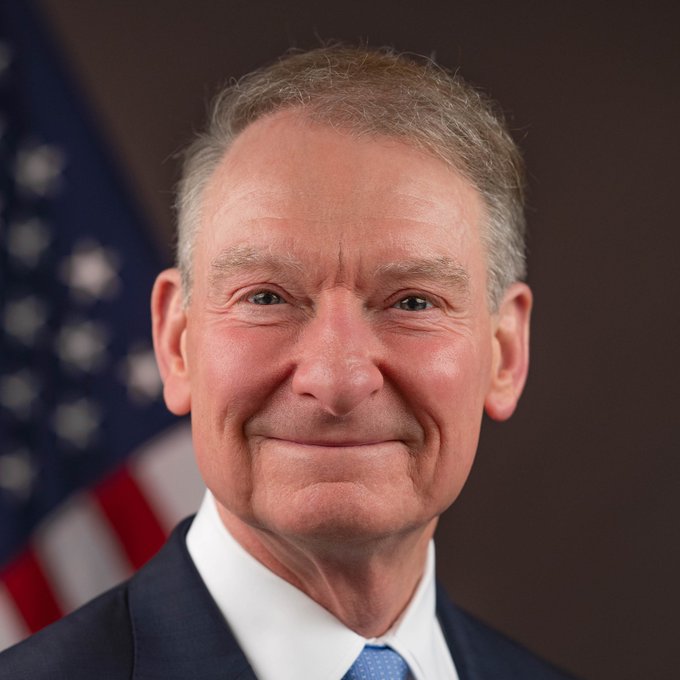 A man in a navy suit and blue tie smiles warmly in an official portrait, with an American flag visible in the background behind him. The image accompanies a post about SEC Chair Paul Atkins signaling forthcoming crypto regulations, suggesting the pictured official is delivering positive news for the crypto industry's regulatory outlook in the US.