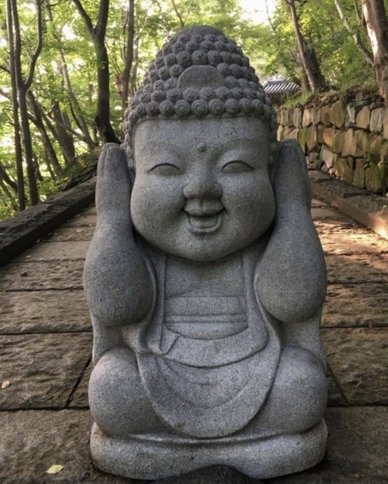 A stone Buddha statue with a smiling face sitting cross-legged outdoors on a wooden path surrounded by green trees and stone walls.
