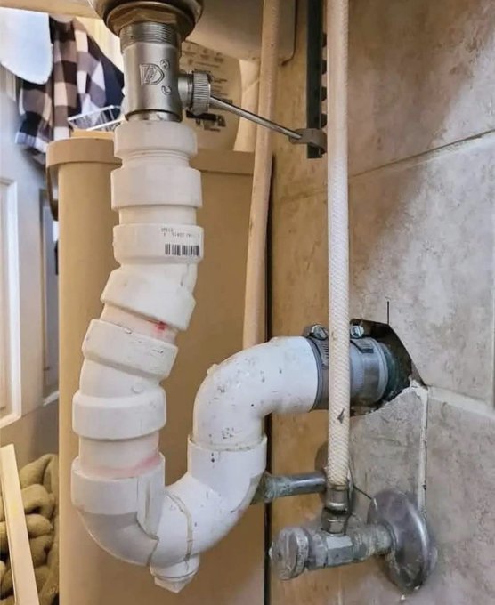 An under-sink view of exposed plumbing featuring white PVC pipes with a P-trap, brass fittings, a chrome valve, and a braided water supply line against a tiled wall.
