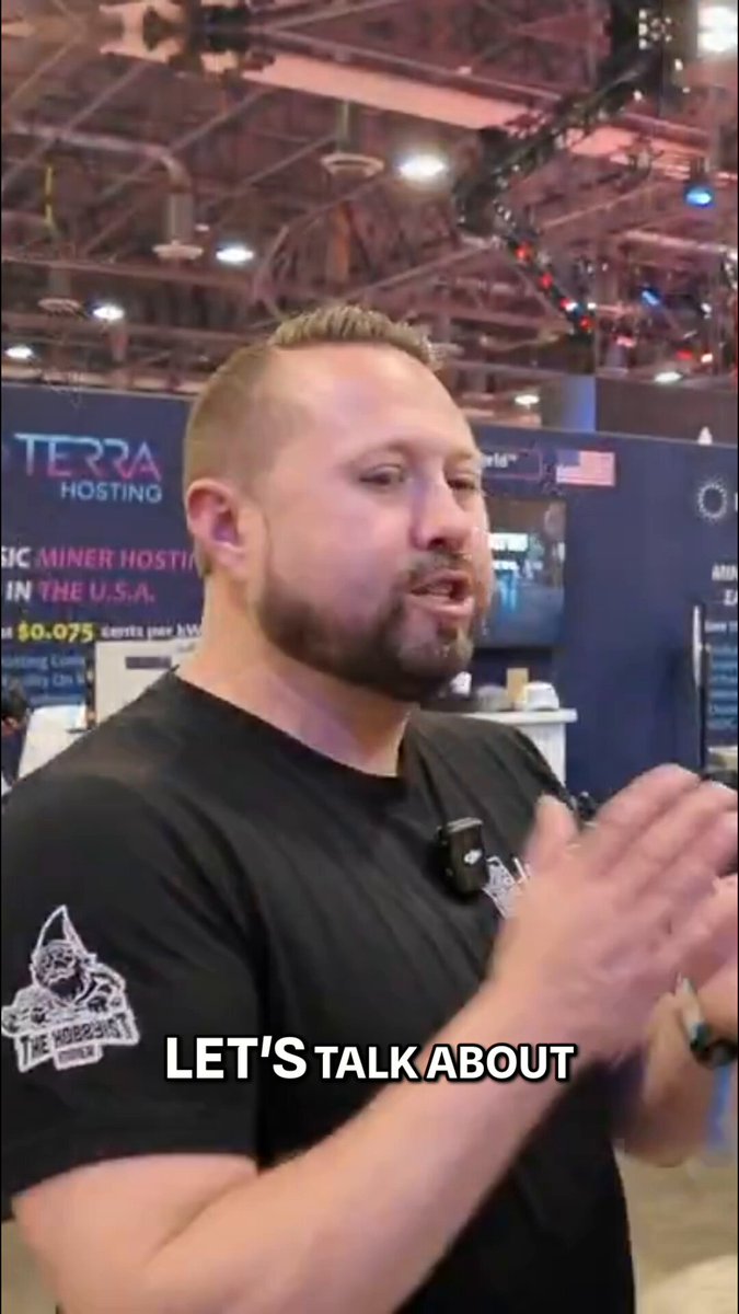 The image shows a man wearing a black shirt, standing in front of a booth at an event or convention. The man appears to be giving a presentation or talking about something related to mining facilities. There are several other people present in the scene as well, indicating that this is likely a large gathering or event.