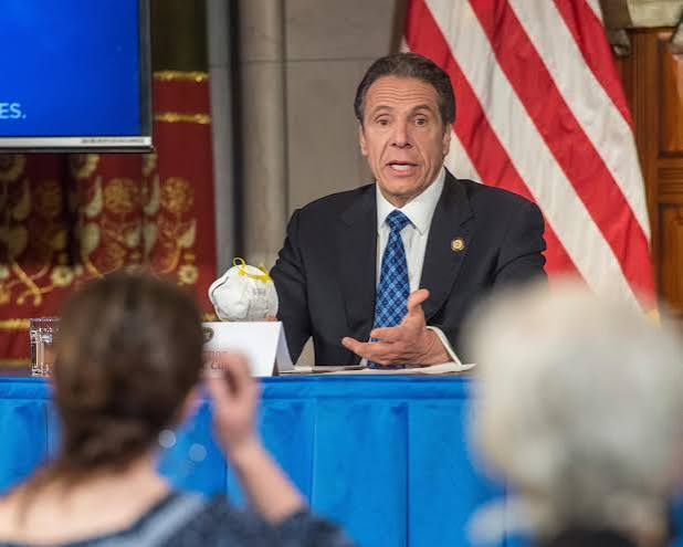 Andrew Cuomo in a suit and blue tie speaking at a press conference with an American flag behind him.