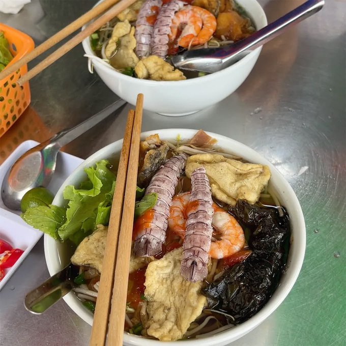 Two bowls of Vietnamese noodle soup (bún) are shown on a metal table, topped with mantis shrimp, shrimp, fried tofu, and herbs, with wooden chopsticks resting across the front bowl.