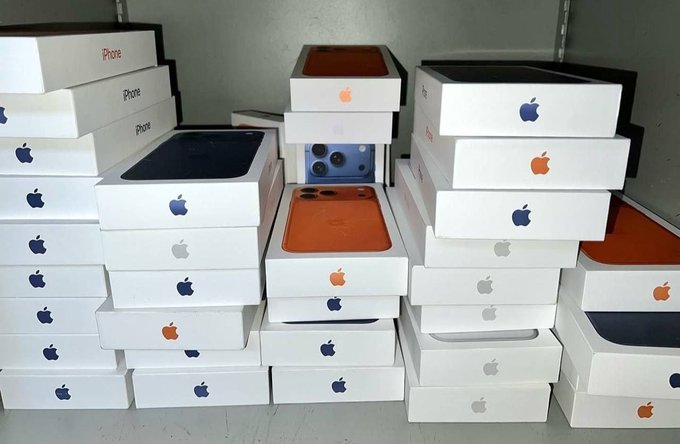 Multiple stacks of white iPhone boxes with Apple logos are piled together, some showing iPhone or iPhone Pro labels, with the boxes displaying images of phones in various colors including orange, blue, black, and white.