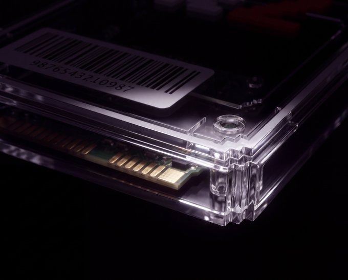A close-up product shot of a transparent acrylic device resembling a card holder with a barcode label (number 9876543210987) and a visible gold connector edge inside.