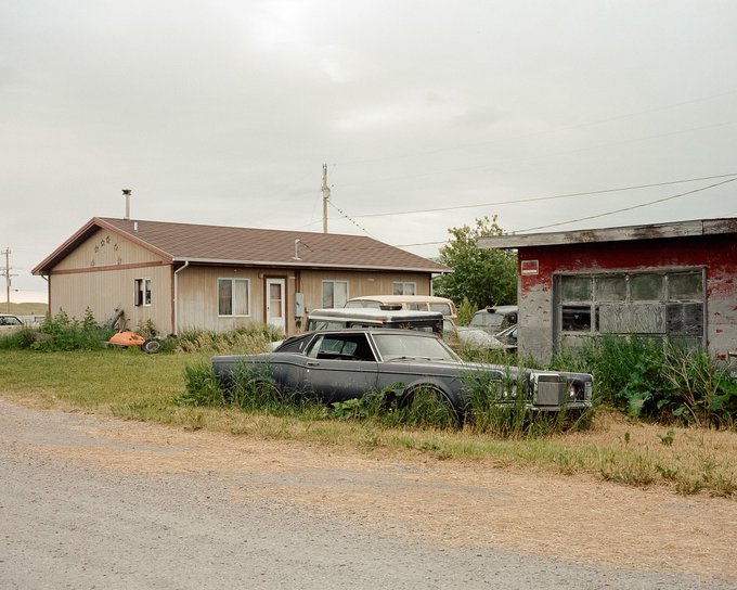 An abandoned gray sedan overgrown with weeds sits in front of a weathered beige house with a brown roof and a dilapidated red shed under an overcast sky.