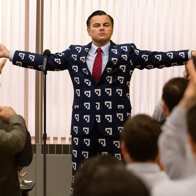 A scene from The Wolf of Wall Street featuring Leonardo DiCaprio in a blue suit covered in white letter 'C' patterns, standing with arms outstretched in front of an applauding crowd.