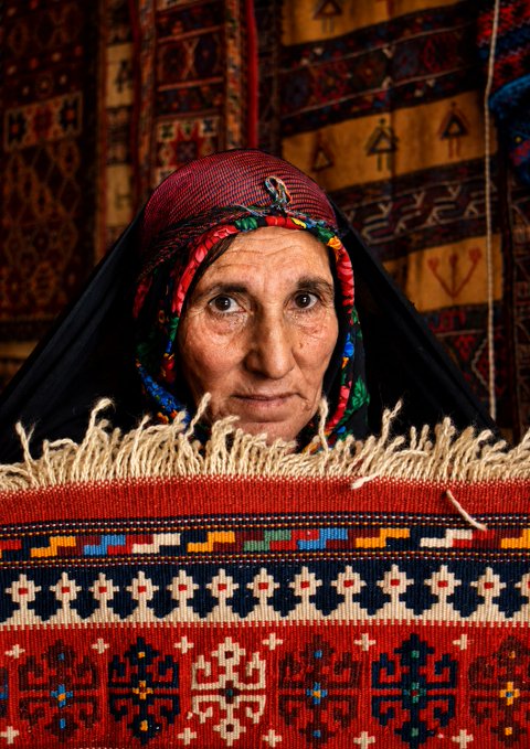 A portrait of an elderly woman wearing a traditional decorated headscarf with colorful beadwork, posed behind a red woven kilim rug featuring geometric and stylized motifs.