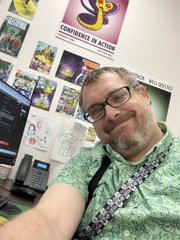 A selfie of a smiling man with glasses in a green patterned shirt, standing in a room with a computer monitor and a wall covered in trading cards including one labeled 'CONFIDENCE IN ACTION'.