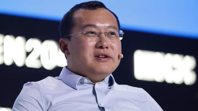 A man with glasses and a headset microphone speaks at what appears to be a financial conference, with partial text "IN20" and "INDEX" visible in the blurred background. The image relates to the post about Star Xu, founder of crypto exchange OKX, who publicly criticized Binance and its CEO CZ regarding the company's failed MiCA license application in Europe.