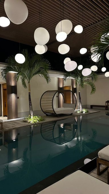 The image shows a large swimming pool with white lights hanging above it, creating an inviting atmosphere for relaxation or social gatherings. The pool area features several palm trees surrounding it, adding a tropical feel to the scene. There are also lounge chairs placed around the poolside, providing comfortable seating options for those who wish to unwind and enjoy the view of the water.