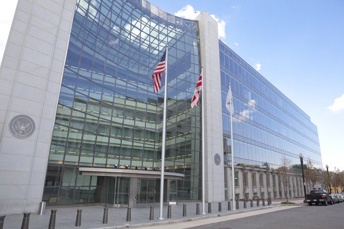 Exterior photograph of a large modern glass office building, likely a U.S. government facility, with American and other flags flying in front.