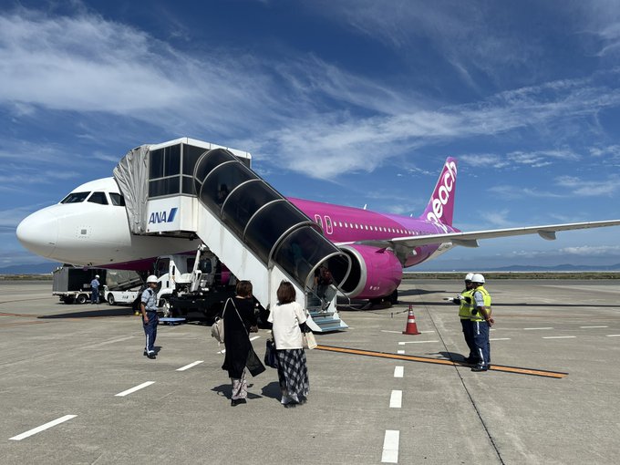 Passengers board a pink and white ANA (All Nippon Airways) "Bee" branded aircraft via a mobile staircase on the tarmac, with ground crew in high-visibility vests assisting at the boarding area under a partly cloudy blue sky—capturing the moment of departure to Sapporo mentioned in the post.