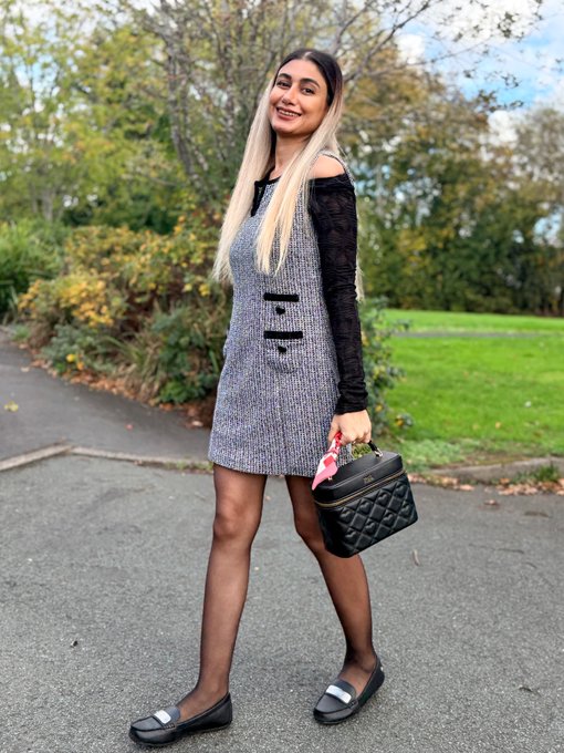A woman with long blonde hair poses outdoors wearing a gray mini dress with black lace sleeves, holding a black quilted handbag and wearing silver ballet flats.
