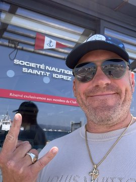 A selfie of a man wearing a black cap, sunglasses, a silver cross necklace, and a white sweater, making a hand gesture in front of a 'SOCIÉTÉ NAUTIQUE SAINT-TROPEZ' banner.