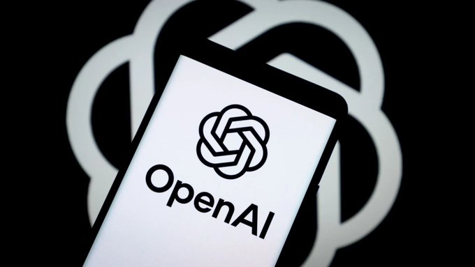 A smartphone displays the OpenAI logo and name on its screen, set against a larger blurred OpenAI emblem in the background, visually representing the company's announcement of its upcoming GPT-5.6 Sol, Terra, and Luna model rollout.