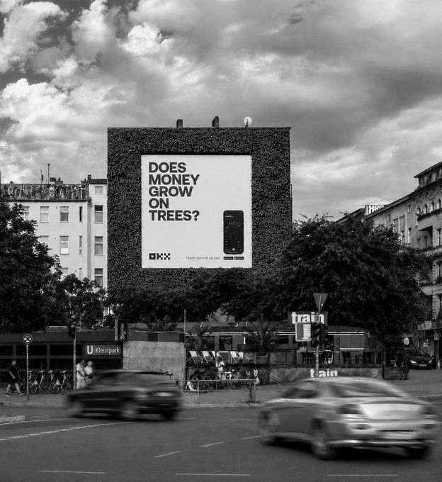 A black-and-white urban scene features a large building-side billboard reading "DOES MONEY GROW ON TREES?" alongside the OKX logo and "TRADE CRYPTO ON OKX," visually tying into the post's claim that the cryptocurrency exchange's marketing presence is hard to miss, even amid blurred passing traffic.