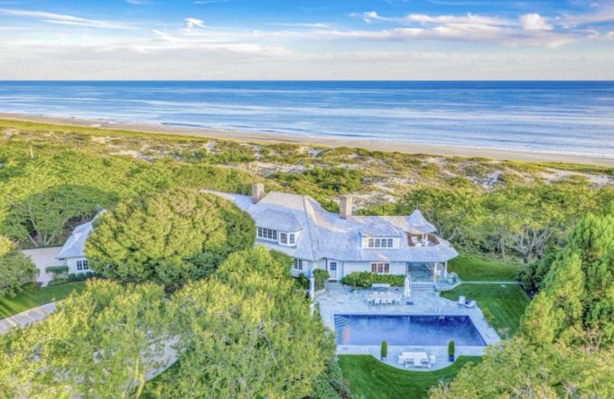 An aerial view shows a luxurious beachfront mansion with a pool and outdoor seating area, surrounded by lush greenery near the ocean.