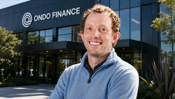 The image features a man standing outside of an office building with the name "Ondo Finance" displayed above him. He has a blue sweater on, giving off a professional appearance. The man appears to be smiling for the camera, possibly posing for a photo or attending an event at the company's location.