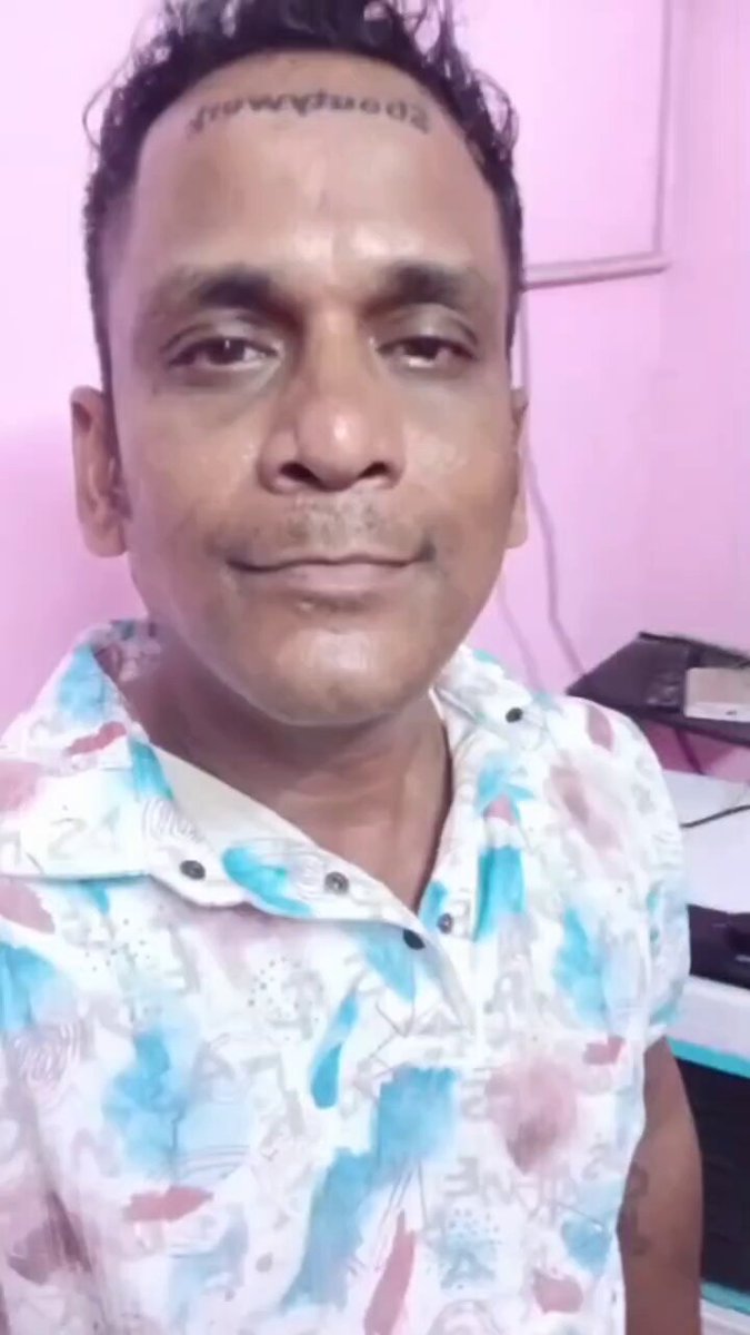 The image features a man wearing a white shirt with blue, pink, and green colors. The man has a tattoo on his forehead that reads "I love you", which adds a personal touch to the photo. The background of the image shows a pink wall, and there is also a computer monitor present in the scene.