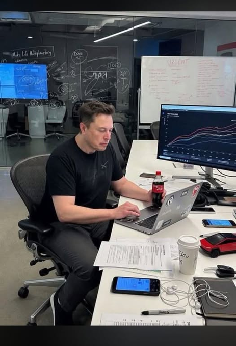 Elon Musk works at a desk with a laptop, surrounded by monitors displaying charts, with a 'URGENT' whiteboard and a Coca-Cola bottle nearby.