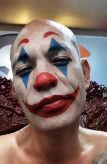 The image features a man with painted clown makeup, including red lips and a blue nose. The man has his shirtless back towards the camera, giving an impression of him posing for the photo. The background includes a chair and a couch, suggesting that this might be taking place in someone's living room or a similar indoor setting.