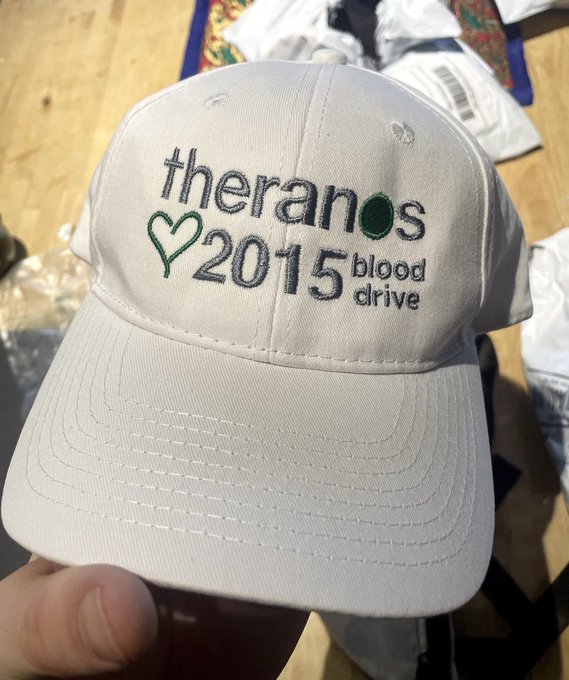 A hand holds a white baseball cap featuring embroidered text that reads 'theranos 2015 blood drive' with a green heart symbol.