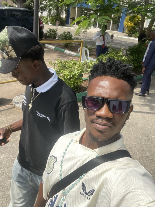 A man wearing sunglasses and a white textured shirt featuring a "BROOKS" eagle logo takes a selfie with a young companion in a black "Balenciaga" shirt, while a person in a business suit stands in the background—capturing a casual Friday moment in Cape Town.