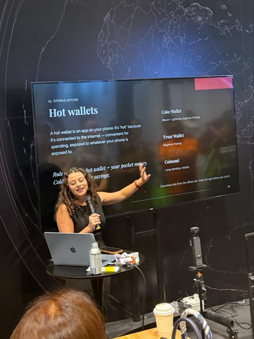A woman presents a slide on "Hot wallets" during a "Storing Bitcoin" educational class, listing options like Cake Wallet, Trust Wallet, and Coloniali as audience members attend the Bitcoin for Beginners session, which the caption notes was so popular the Q&A ran 15 minutes over time.