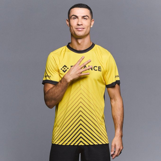 A man wearing a yellow and black "BINANCE" branded jersey poses against a gray background, with his hand placed on his chest, aligning with the post's celebration of him in Binance colors alongside the caption "The GOAT in #Binance colors Binancians everywhere."