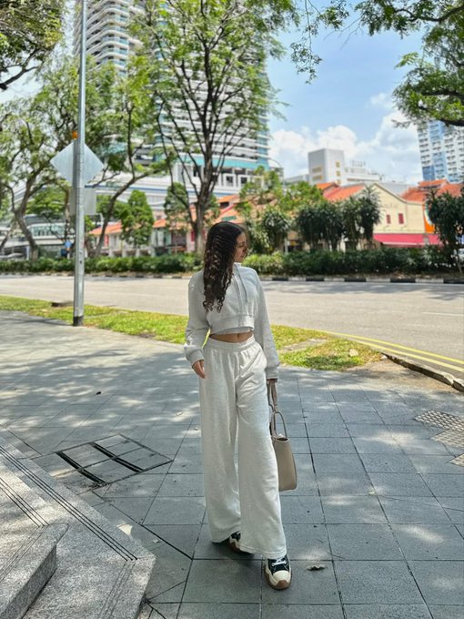 A woman in a white cropped jacket and pants stands on a sidewalk looking away, with trees and buildings in the background.