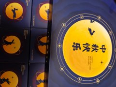 A stack of elegant dark blue gift boxes featuring a golden full moon design with the Chinese characters "胡家洪宇" (Hu Jiā Hóng Yǔ) and a bat silhouette, presented as a Mid-Autumn Festival gift set from Envo. The packaging's celestial moon imagery ties directly to the festival theme, complementing the post's appreciation for the mooncakes and branded peripherals (hats and shirts) included in the promotional package.