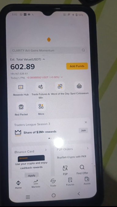 A photo of a smartphone displaying the Binance app home screen, showing an estimated total value of 602.89 USDT, a Rewards Hub, P2P orders, and a Traders League Season 3 promotion.