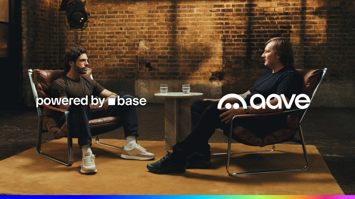 Two men sit facing each other in leather chairs against a brick-wall backdrop, with a small table and water glasses between them. White logos read “powered by base” on the left and “aave” on the right, matching the post about Aave bringing lending and borrowing to tokenized stocks on Base.
