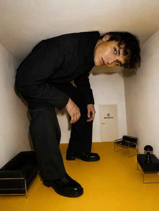 A young man in a black outfit crouches in a stylized room with a yellow floor, with a small Binance-branded sign visible on the door.