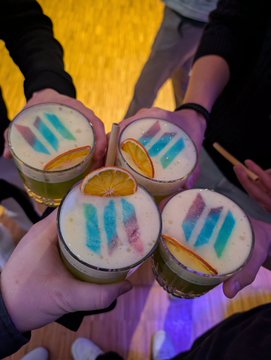 The image shows four people holding glasses of colorful drinks, possibly cocktails or smoothies, with a variety of colors including blue, green, orange, and yellow. The drinks are garnished with slices of oranges and other fruits. A person's hand is visible in the foreground, while another person can be seen further back on the right side of the image. The background appears to be a room where people have gathered for an after-party or celebration.