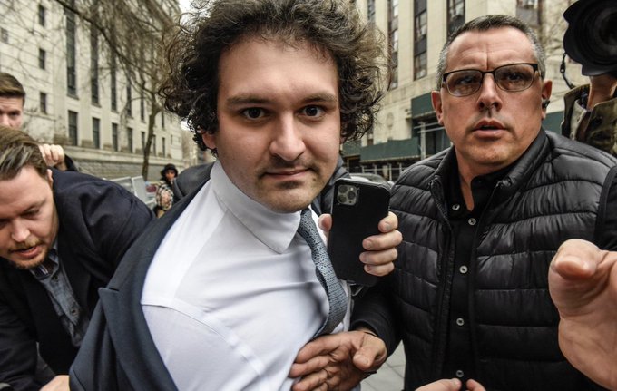 A curly-haired man in a white shirt and tie is escorted through a crowd outside a building, holding a phone and being guided by a man in a black puffer vest and glasses.